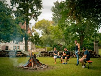 Vier Männer entspannen sich um ein Lagerfeuer auf einer Wiese. Im Hintergrund sind Bäume und ein Haus zu sehen.